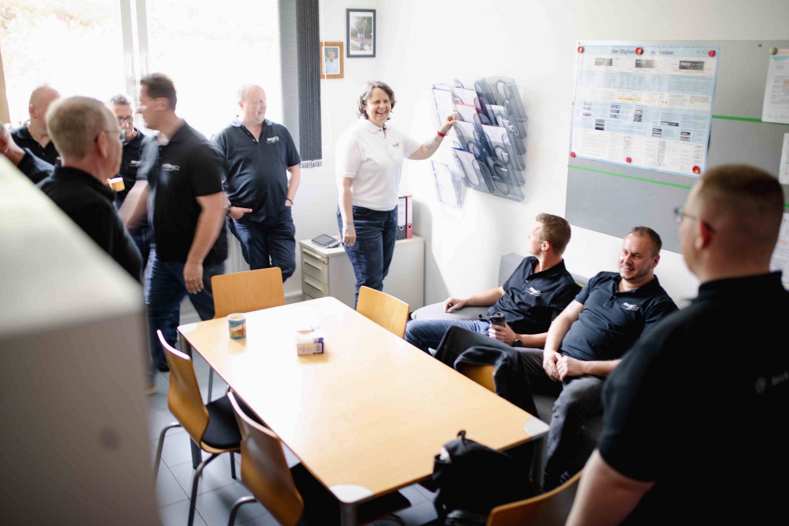 Größere Gruppe sitzt und steht in einem Aufenthaltsraum, lacht und sie unterhalten sich.